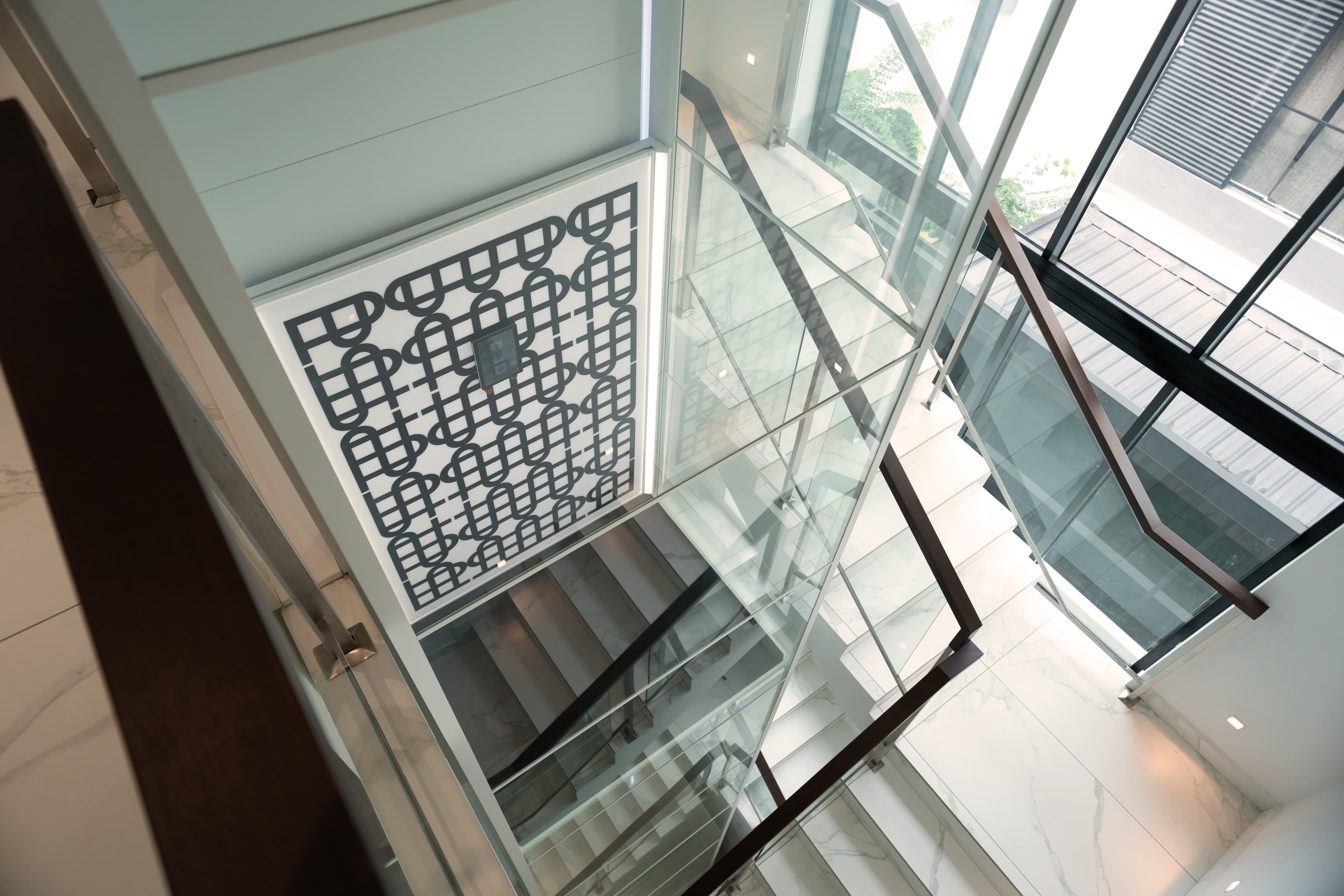 Looking down a glass lift shaft that runs beside a white marble staircase with timber handrails and a patterned feature screen