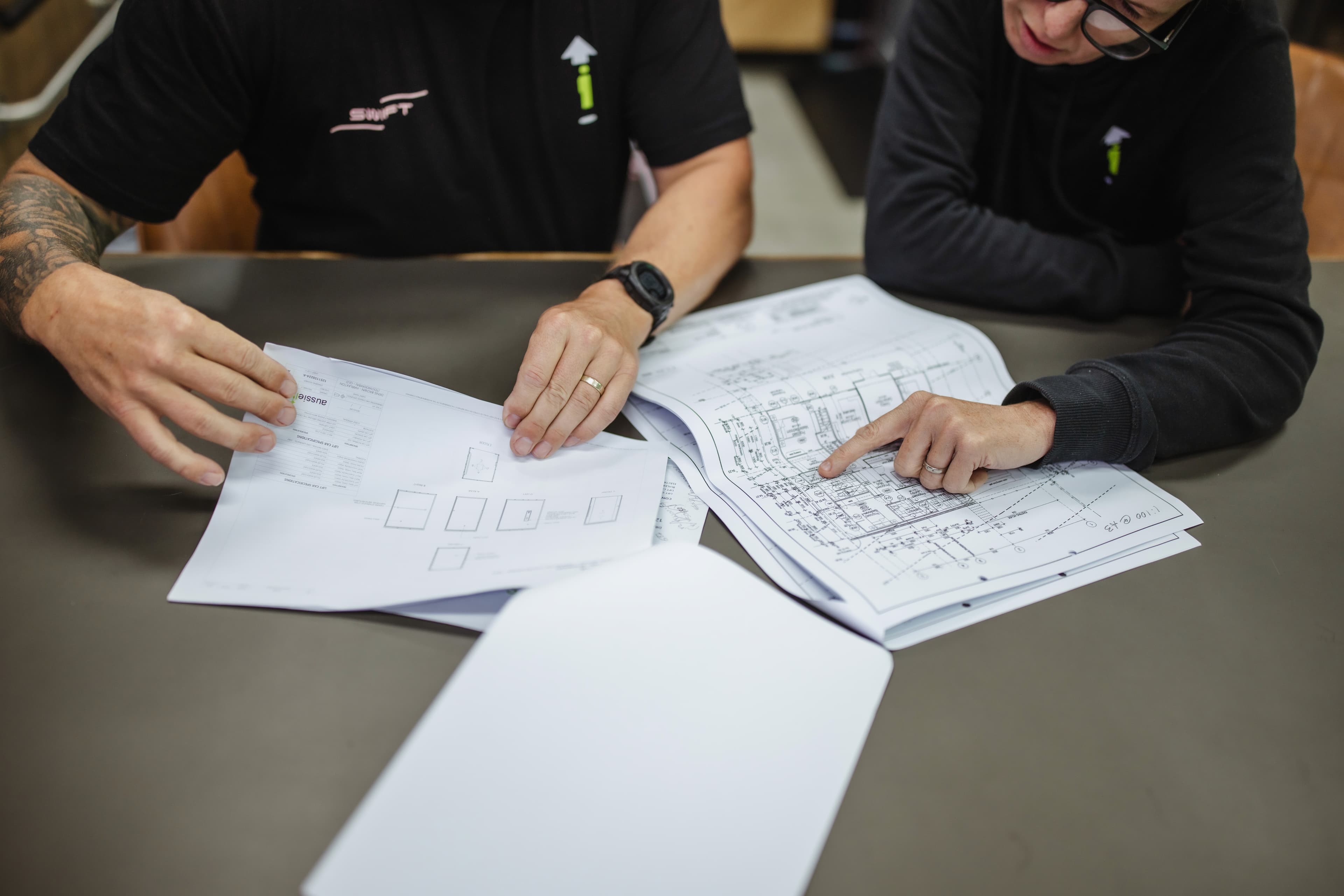 Close-up of two people pointing at printed lift layout drawings spread on a table