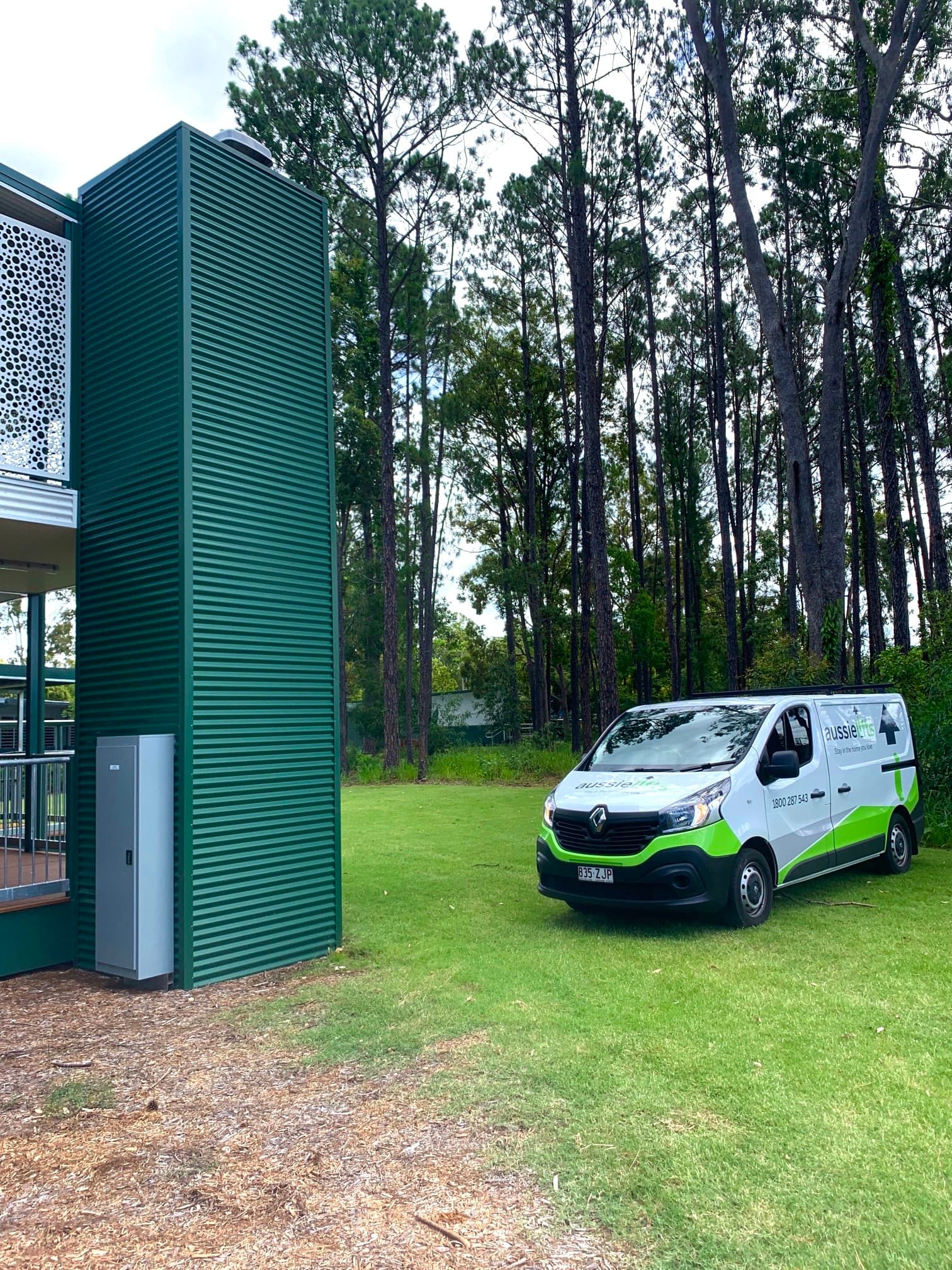 Green clad external lift shaft beside a raised deck, with a white Aussie Lifts van parked on the lawn under tall pine trees
