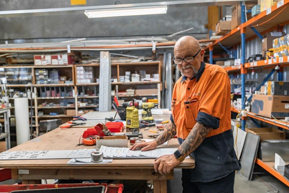 Man in an orange work shirt and glasses smiling at a workbench with tools and drawings, shelves of parts behind him