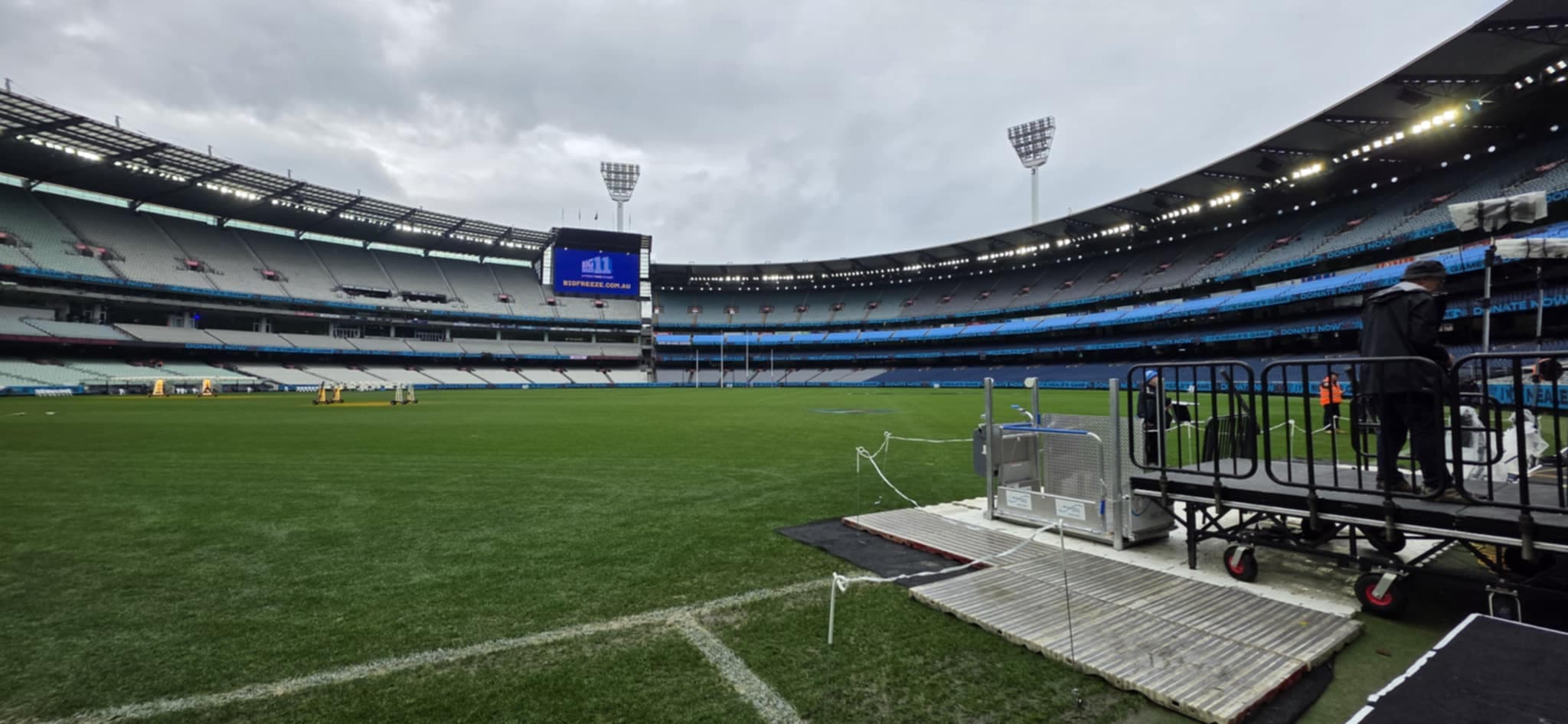 Portable platform lift and ramp set up at the edge of the grass in a large empty sports stadium