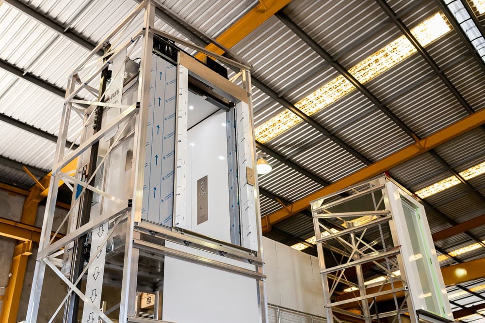 Lift car with its door open inside an aluminium shaft frame under assembly in a factory, a second glass shaft frame beside it