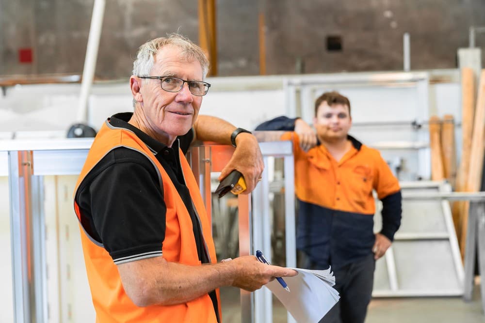 Older man in an orange hi-vis vest holding papers and smiling in a workshop, a younger worker in an orange shirt behind him