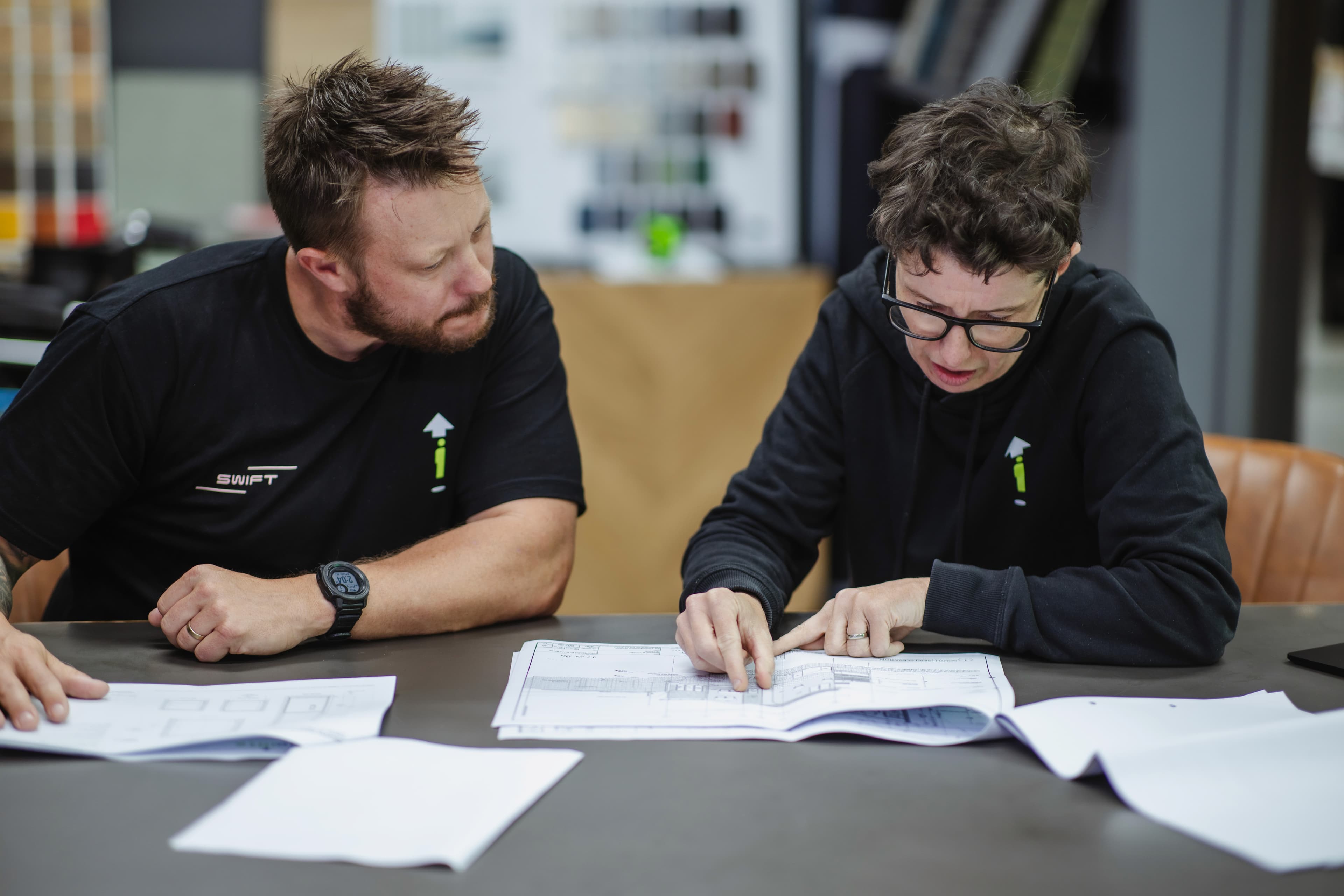 Two Aussie Lifts staff at a table reviewing printed lift plans, finish samples on the wall behind them