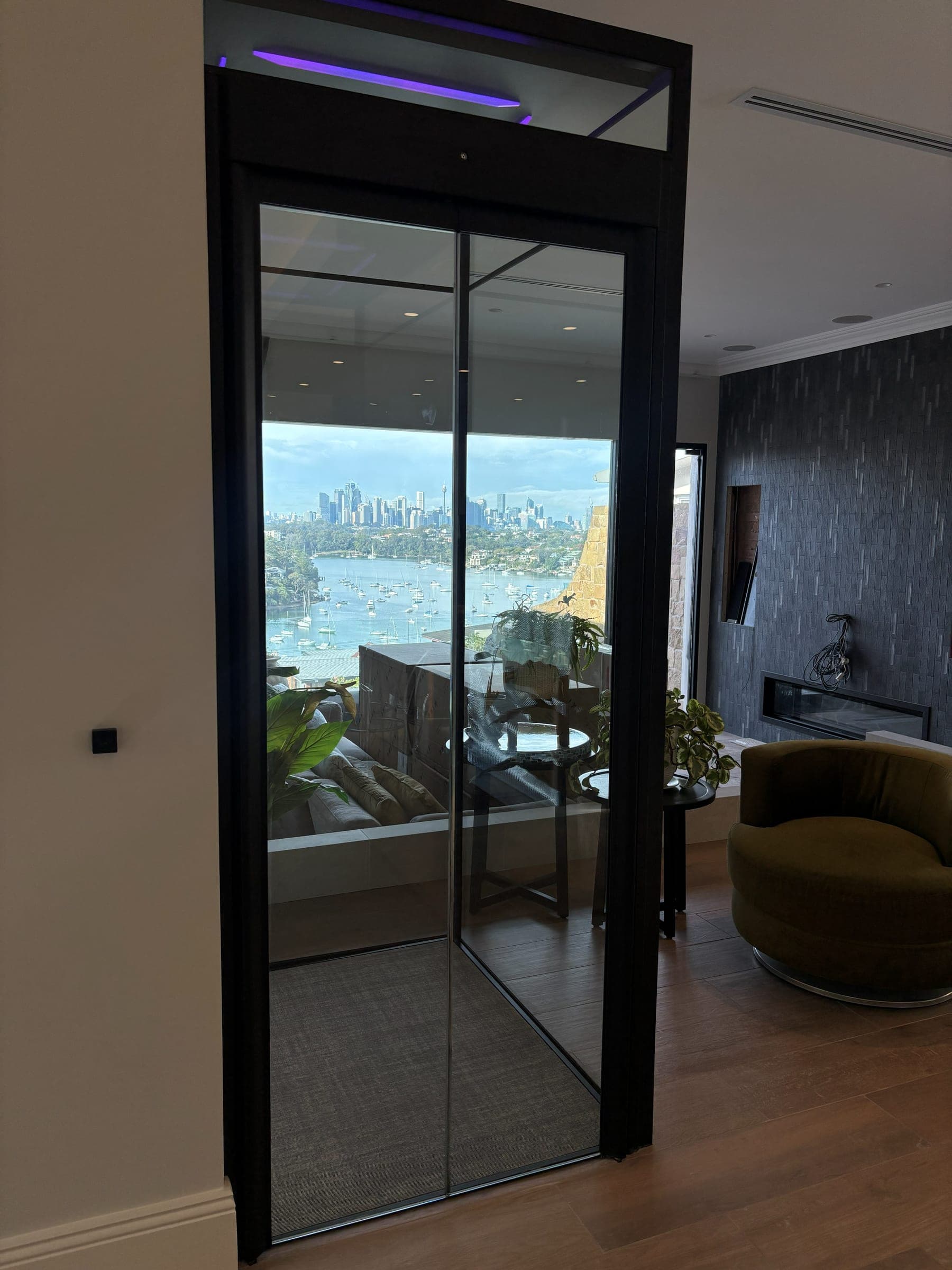 A Swift Pro home lift with a full glass shaft in a Longueville home, the Sydney city skyline visible straight through it across the water